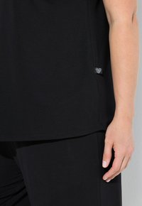 Zwart katoenen T-shirt met een relaxed fit, ronde zoom en een klein merkembleem aan de zijkant. Close-up toont een hand die op de stof rust.