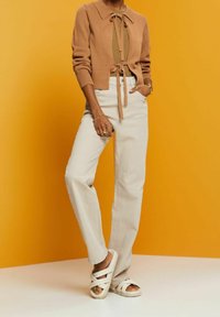 Personne portant un cardigan beige sur un haut olive, jean blanc taille haute, sandales blanches, debout devant un fond jaune.