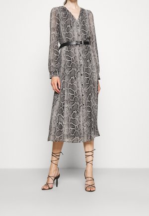 Femme portant une robe midi grise à imprimé serpent avec manches longues, ceinture noire, et sandales à talons hauts noires à brides sur un fond uni.