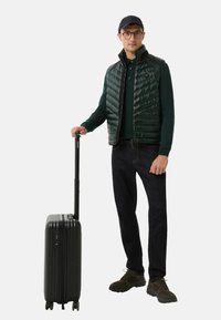 Mann in dunkelgrünem gesteppter Weste, Mütze, Brille und dunkler Jeans steht und hält einen schwarzen Rollkoffer am Griff.
