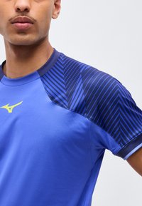 Άνδρας με μπλε αθλητική μπλούζα με σκούρες μπλε λωρίδες σε σχήμα βέλους στους ώμους και κίτρινο λογότυπο στο στήθος, σε ανοιχτό φόντο.