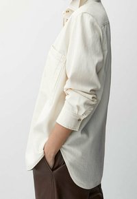 Camicia color crema a maniche lunghe realizzata in tessuto morbido, con taschino sul petto e polsini con bottoni, mostrata di lato per evidenziare il taglio e la lunghezza.