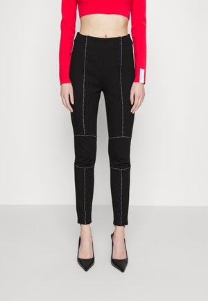 Pantalon noir ajusté avec des coutures blanches contrastantes, jambes fuselées et ceinture élastique, associé à un haut rouge court à manches longues et des talons noirs.