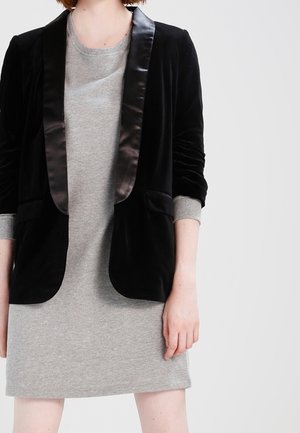 Person iført en grå langærmet kjole og en sort fløjlsblazer med satinrevers, stående mod en ensfarvet lys baggrund.