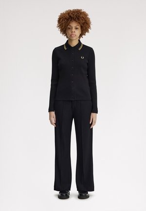 Frau steht frontseitig, trägt ein schwarzes langärmliges Poloshirt mit gelben Streifen am Kragen und schwarze Hosen mit weitem Bein.