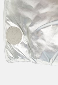 Zilveren jack met een glanzende, metallic afwerking, voorzien van een ronde, crème-kleurige "Save the Duck" logo patch en een gequiltde textuur.