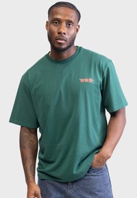 Grünes Baumwoll-T-Shirt mit kurzen Ärmeln und Rundhalsausschnitt, mit einem kleinen roten Druck auf der linken Brustseite. Getragen mit Jeansshorts.