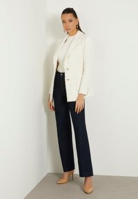 Blazer bianco con texture e bottoni dorati, top bianco a costine, jeans scuri e décolleté color nude. In piedi contro uno sfondo di parete chiara.