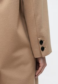 Bézs színű gyapjúkabát ujja sima textúrával és három fekete gombbal a mandzsettán, precíz varrási részletekkel.