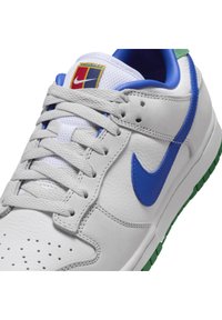 Vita lädersneakers med blå Nike-swoosh, grå snören, grön sula och en flerfärgad logotypapplikation på den vita mesh-tungan.