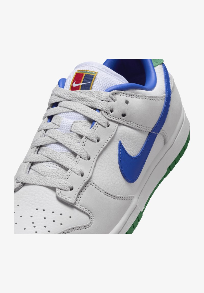 Vita lädersneakers med blå Nike-swoosh, grå snören, grön sula och en flerfärgad logotypapplikation på den vita mesh-tungan.