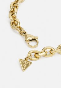 Bracciale in oro con grandi anelli intrecciati. Presenta un gancio a forma di aragosta e un ciondolo triangolare con la scritta "GUESS" impressa al centro.