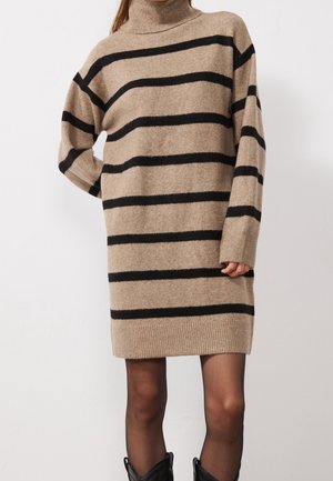 Kvinde iført beige strikket kjole med sorte vandrette striber, højt halsrør, lange ærmer, gennemsigtige sorte strømpebukser og sorte støvler.