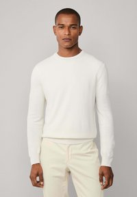 Muškarac u bijelom puloveru s dugim rukavima i svijetlosmešavim hlačama, stoji nasuprot jednostavne svijetlosive pozadine.