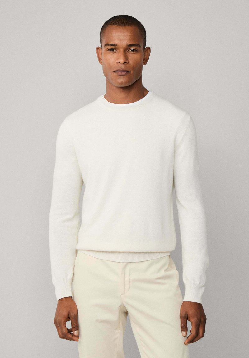 Muškarac u bijelom puloveru s dugim rukavima i svijetlosmešavim hlačama, stoji nasuprot jednostavne svijetlosive pozadine.