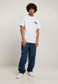 T-shirt di cotone bianco con un design grafico nero sul petto, abbinato a jeans in denim blu a taglio largo e scarpe da ginnastica bianche.