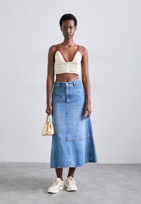 Top corto all'uncinetto di colore crema con dettagli rossi, abbinato a una gonna maxi in denim blu chiaro. Presenta una borsa elegante e sneakers bianche.