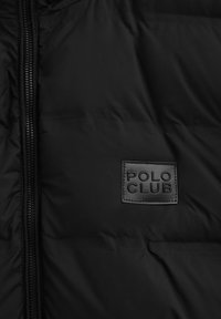 Svart vadderad jacka med framdragkedja och en sydd fyrkantig lapp märkt "POLO CLUB" på bröstområdet.