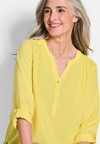 Blusa gialla, leggera, con scollatura a V, sette bottoni frontali e maniche arrotolate. Texture liscia e vestibilità rilassata.