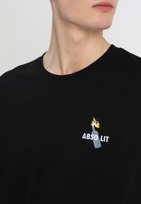 Černé bavlněné tričko s vyšitým designem lahve plné plamenů a textem „ABSOLIT“ v bílé barvě. Kulatý výstřih.