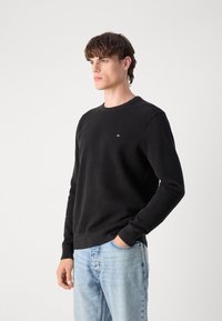 Zwart gebreid sweatshirt met een gestructureerd patroon, ronde hals en een klein logo op de borst. Gecombineerd met lichtblauwe spijkerbroek.