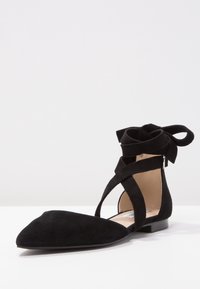 Scarpa nera in suede a punta con cinturini incrociati alla caviglia e fiocco legato sul retro, posta su uno sfondo bianco.