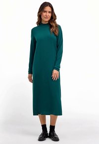 Langärmeliges, smaragdgrünes Kleid mit hohem Ausschnitt, knielang, weicher Textur und schlichter Silhouette; kombiniert mit schwarzen Schuhen.