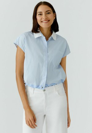 Lachende vrouw met donker schouderlang haar draagt een lichtblauwe blouse met korte mouwen en witte strepen, een witte kraag en witte broek tegen een effen achtergrond.