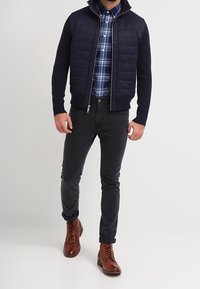 Námořnická prošívaná bunda s vysokým límcem, zkombinovaná s modrou kostkovanou košilí, černými slim-fit kalhotami a hnědými šněrovacími botami.