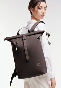 Hnědý batoh s rolovacím vrškem vyrobený z odolného materiálu, se nastavitelnými ramenními popruhy, přední kapsou na zip a logem.