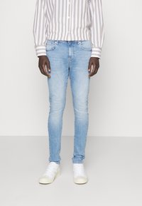 Jeans ajustados de un azul claro con cinco bolsillos, un ligero desvanecimiento y un poco de elasticidad. Combinados con una camisa de rayas de manga larga y zapatillas blancas.
