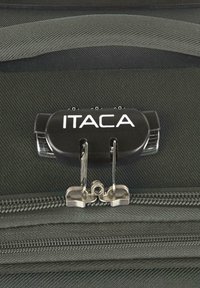 Candado negro para equipaje con el texto "ITACA", dos cremalleras metálicas y fondo de nailon texturizado. El candado tiene un dial de combinación.