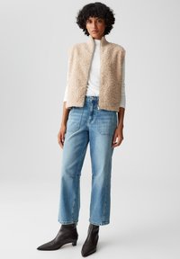 Gilet beige senza maniche in tessuto testurizzato, indossato sopra una maglietta bianca a maniche lunghe, abbinato a jeans larghi blu chiaro e stivali neri alla caviglia.