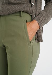 Pantaloni su misura verde oliva con tasche laterali, abbinati a un cardigan in maglia marrone con polsini a coste e bottoni.