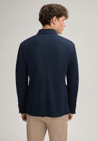 Marineblauer Blazer aus strukturiertem Stoff, mit Reverskragen, geradem Schnitt und zwei seitlichen Taschen, sowie Knopfdetails an den Ärmeln.