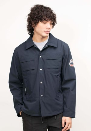 Uomo con capelli ricci che indossa una giacca blu navy abbottonata con tasche sul petto e una toppa sulla manica sinistra, che guarda verso il basso a destra.