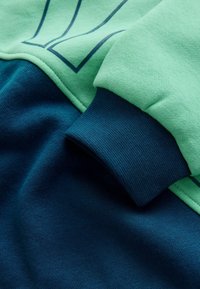 Materiał bluzy w kolorze zielonym i granatowym, z zieloną górną częścią oraz niebieskimi akcentami i teksturowanym mankietem. Gładka tekstura i spójna kolorystyka.