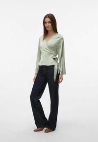 Ljusgrön satinblus med lång ärm, knuten i midjan, i kombination med mörkblå vida jeans och bruna platta skor.
