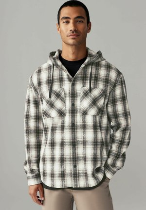 Plaid flannel shirt in black and white with a hood, featuring two front pockets and adjustable drawstrings. Soft texture, relaxed fit.