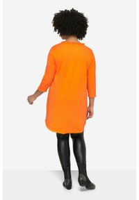 Orange Tunika mit Dreiviertelärmeln, abgerundetem Saum und ausgefranster Halslinie, kombiniert mit schwarzen, glänzenden Leggings und beschlagenen Schuhen.