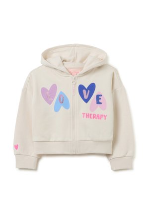 Felpa corta color crema con zip frontale, caratterizzata da vivaci disegni di cuori e la scritta "LOVE THERAPY." Accento a forma di cuore rosa sulla manica.