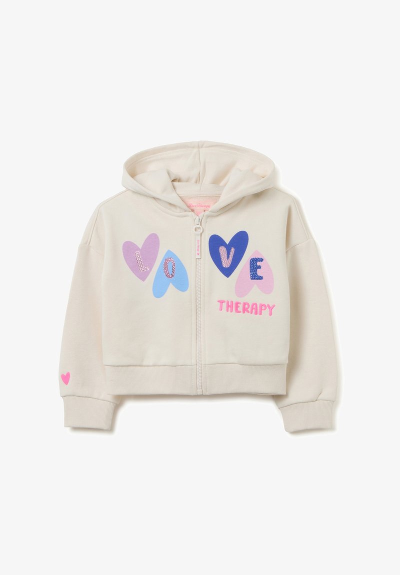 Gekürzte cremefarbene Hoodie mit einem Frontreißverschluss, versehen mit bunten Herzdesigns und dem Text "LOVE THERAPY." Rosa Herzakzent am Ärmel.