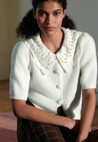 Hvid ribbet cardigan med en tekstureret, blonderkrave. Har korte ærmer og tre knapper foran. Modellen sidder op ad en neutral baggrund.