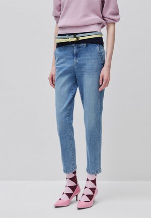 Persona vistiendo vaqueros azul claro, calcetines rosa y burdeos con diseño rombos, tacones altos rosas, cinturones amarillo y azul claro, y un suéter rosa con mangas remangadas.