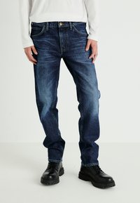 Jeans de mezclilla azul oscuro con un acabado desgastado, diseño de pierna recta, cinco bolsillos y cierre de botón, combinados con botas negras con cordones.