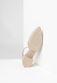 Scarpa con tacco alto beige, punta affusolata, cinturino trasparente e suola testurizzata. Presenta un marchio italiano sul fondo.