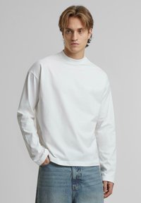 Langärmliges weißes Baumwollhemd mit hohem Kragen und lockerer Passform. Dezentes Logo auf der Brust. Getragen mit hellblauer Jeans.