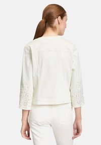 Biała kurtka dżinsowa z rękawami o długości trzech czwartek, ozdobiona koronkowymi wycięciami i drobnymi zdobieniami na rękawach; gładka tekstura.