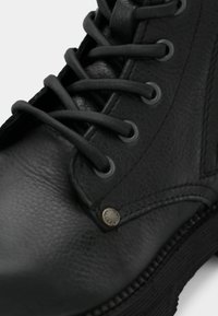 Botte en cuir noir avec une texture lisse, des lacets ronds, des oeillets en métal et un accent métallique rond près de la base.