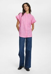 Camicia rosa a maniche corte con ricami floreali, chiusura con bottoni e maniche a balze. Abbinata a jeans a gamba larga blu e scarpe nere.